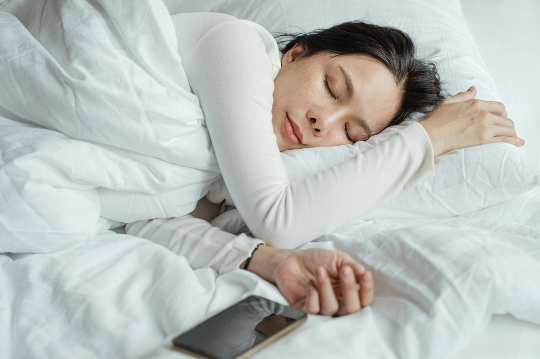Peaceful woman sleeping serenely in crisp white luxury sleepwear on soft white bedding, embodying the ultimate comfort and restful elegance of premium nightwear, with smartphone nearby on the bed.